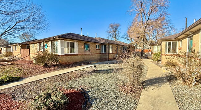 Encantadores apartamentos de ladrillo de una sola planta rodeados de exuberantes jardines y senderos arbolados bajo un cielo azul claro. - Flats at 16th