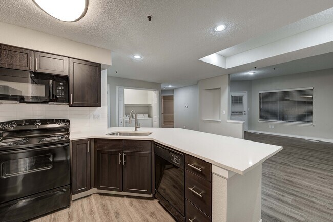 Modern kitchen featuring sleek dark cabinetry, spacious countertops, and stainless steel appliances seamlessly leading into an open-concept living area with stylish flooring.