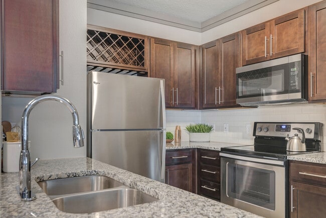 Cocina moderna con gabinetes de madera, salpicadero de azulejos blancos, encimeras de granito y electrodomésticos de acero inoxidable - Avalon Mooresville Apartamentos