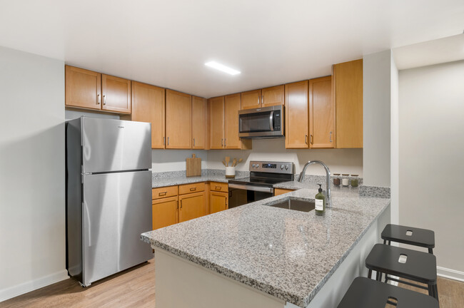 Cocina con acabados actualizados con encimeras de granito moteado de gris, gabinetes de roble, electrodomésticos de acero inoxidable y pisos de superficie dura. - Kanso Silver Spring Apartamentos