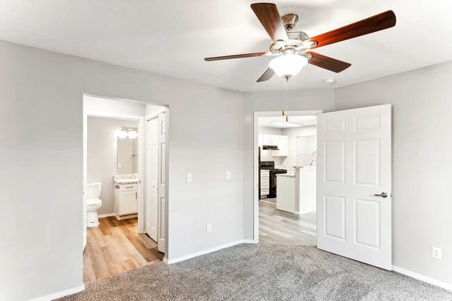 Bedroom with carpet flooring, ceiling fan with light, neutral walls, and open doorways leading to bathroom and kitchen area