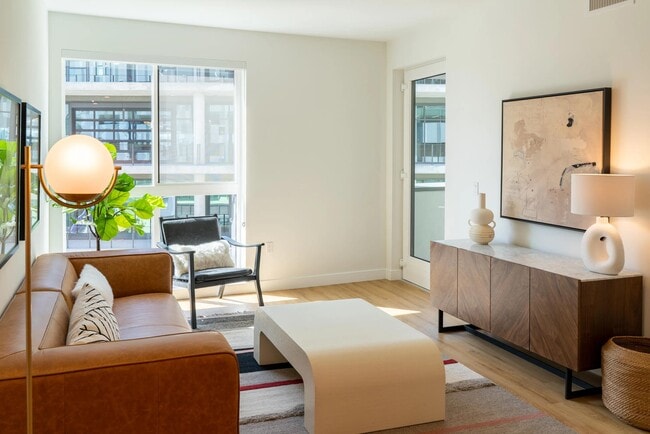 Living area with floor-to-ceiling windows, warm wood-style plank flooring, and a private balcony that brings in natural light and neighborhood views.