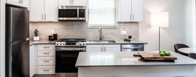 Design Collection II (South Building) Kitchen with white cabinetry, gold pulls, beige tile backsplash, grey quartz countertops, stainless steel appliances, and hard surface flooring - Avalon Somerville Station Apartments