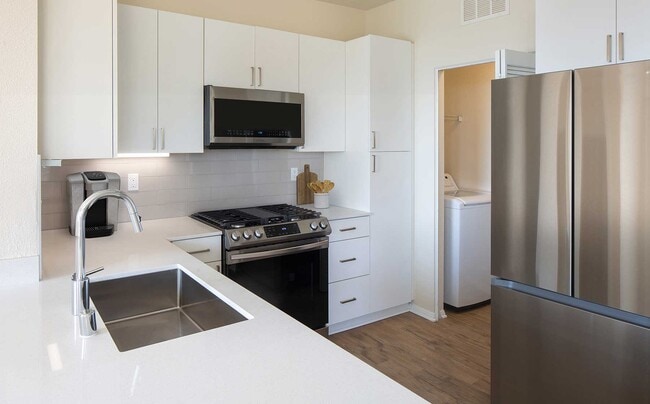 Renovated Package II kitchen featuring white cabinetry, white quartz countertops, stainless steel appliances, and hard surface flooring. In-unit laundry with full sized washer and dryer