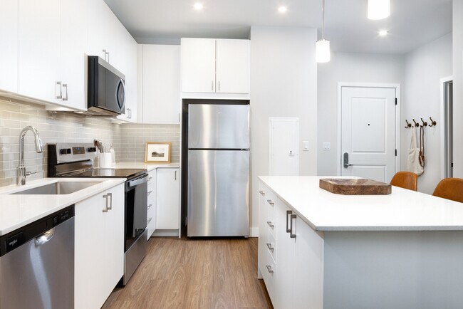 Design Collection I kitchen with stainless steel appliances, white cabinetry, grey tile backsplash, under-cabinet lighting, white quartz countertops, and hard surface flooring - Avalon Quincy Adams