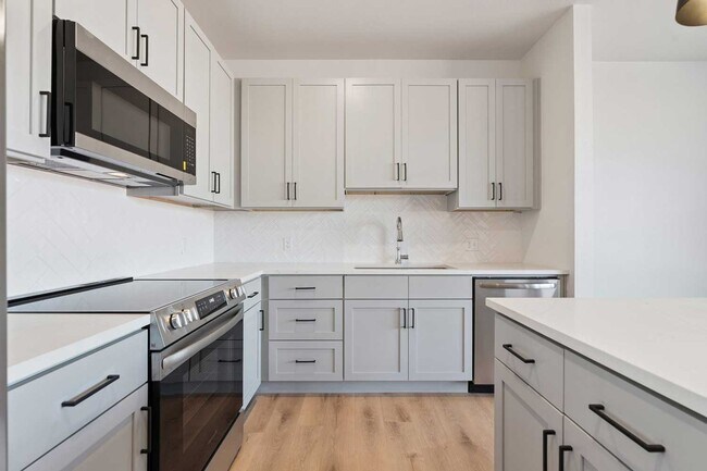 Modern kitchen with gray shaker cabinets, quartz countertops, stainless steel appliances, and herringbone tile backsplash