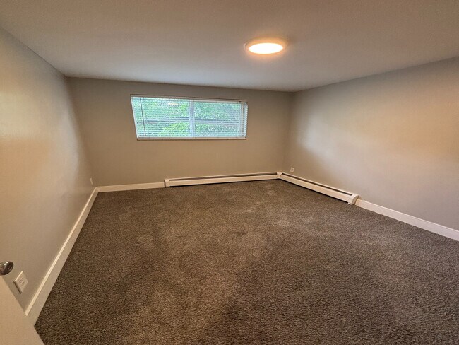 Cozy room with soft carpet flooring and a bright window, perfect for personalization and comfort. - Copperwood Apartments