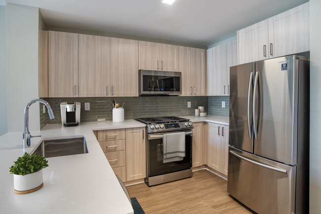 Renovated Package II kitchen with oak cabinetry, white quartz countertops, stainless steel appliances, grey title backsplash, and hard surface flooring - Avalon at Gallery Place Apartments