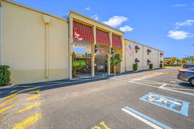 Inviting apartment entrance with vibrant red accents, American and state flags, lush greenery, and ample parking.