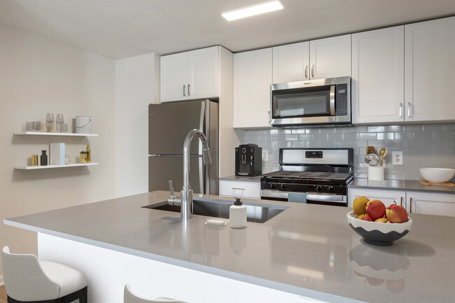 Finish Package II kitchen with white cabinetry, grey quartz countertops, extended hard surface flooring in select units, stainless steel appliances, and tile backsplash - Avalon Westbury Apartments