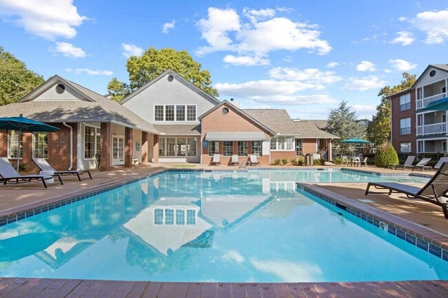 Community pool area featuring lounge chairs, umbrellas, and brick clubhouse under a bright blue sky with trees