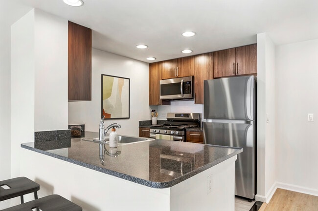 Updated Finishes kitchen with espresso cabinetry, dark speckled granite countertops, white tile backsplash, stainless steel appliances, and hard surface flooring - AVA Fort Greene Apartments