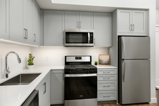 Apartments con modernas cocinas con gabinetes de color gris, blanco, encimeras de piedra de cuarzo, placa para salpicaduras de azulejos blancos, electrodomésticos de acero inoxidable, y pisos de superficie dura