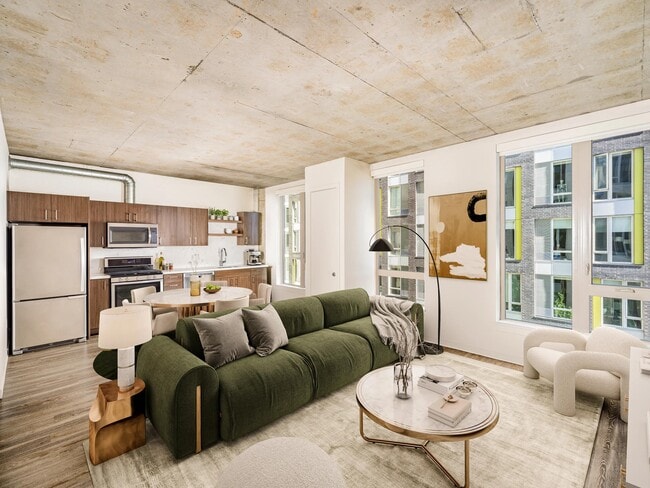 An open apartment home with concrete ceilings, expansive windows, and a streamlined kitchen, offering flexible living space filled with natural light.
