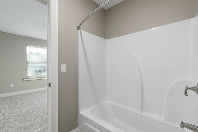 Bright and modern bathroom with elegant white bathtub and sleek fixtures, featuring a cozy adjoining carpeted bedroom with a large window.