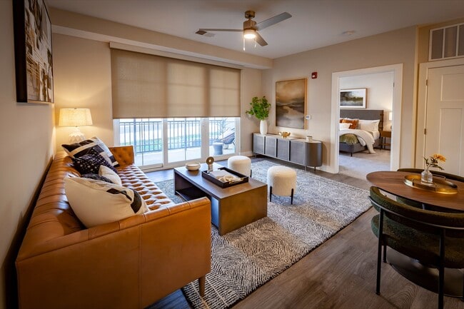 Elegant apartment living room with a chic leather sofa, modern decor, abundant natural light, and an open layout, including dining area, flowing into a cozy bedroom.