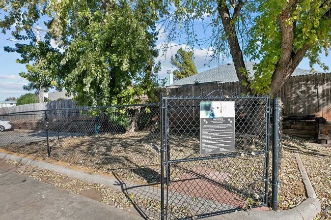 Fenced dog park entrance with chain-link gate, bark park signage, gravel ground cover, and mature shade trees