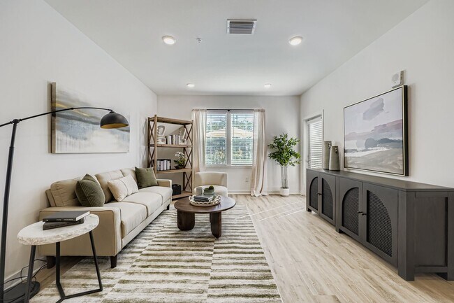 Bright and modern living room featuring a cozy beige sofa, stylish artwork, and abundant natural light from large windows. - Magnolia at Palm Bay