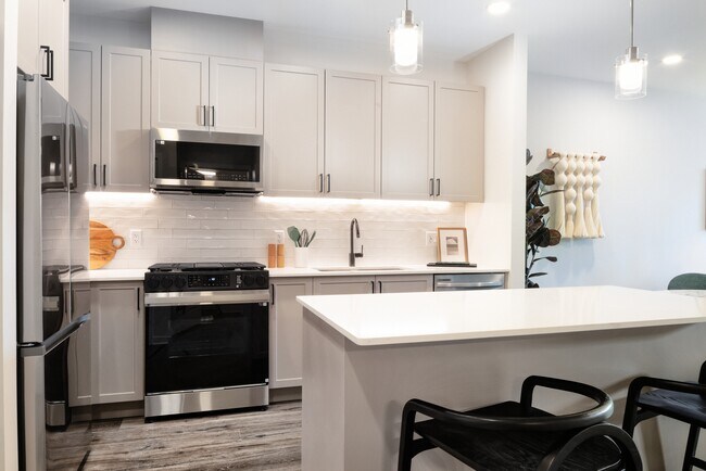 Modern kitchen with grey cabinetry, white tile backsplash, white quartz countertops, stainless steel appliances, and pendant lighting - Avalon Princeton on Harrison Apartments