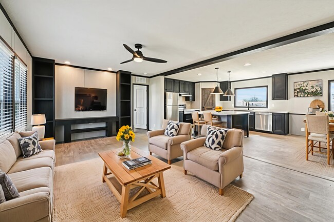 Sala de estar abierta con sofás beige, ventilador de techo negro y cocina con iluminación colgante y zona de comedor.