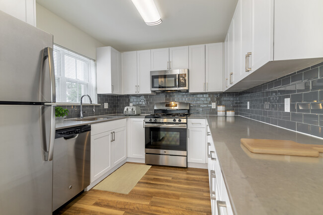 Renovated Package II kitchen with white cabinetry, grey quartz countertops, grey tile backsplash, stainless steel appliances, and hard surface plank flooring throughout common areas. Soft-close cabinets and microwaves available in select homes - eaves Wilmington and Wilmington West Apartments