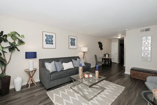 Spacious living room with dark hardwood floors, gray sofa, glass coffee table, framed blue artwork, and indoor tree