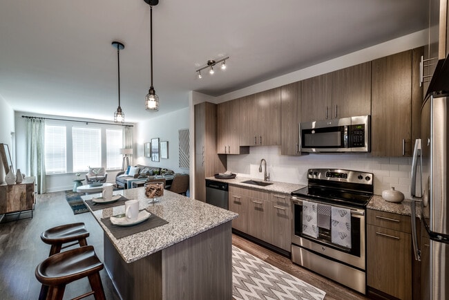 Open concept kitchen with brown cabinetry, granite countertops, kitchen island, stainless steel appliances, tile backsplash and hard surface flooring - Avalon Arundel Crossing Apartments