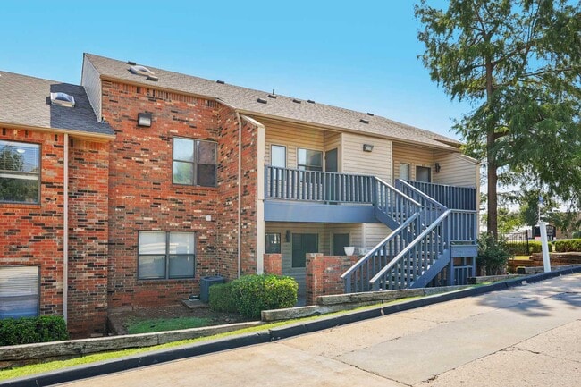 Two-story apartment building exterior with red brick facade, blue staircase, upper balcony, shrubs, and tall trees
