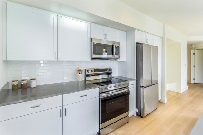 Renovated Package I kitchen with white cabinetry, grey quartz countertops, white subway tile backsplash, stainless steel appliances, and hard surface flooring - Avalon Towers Bellevue