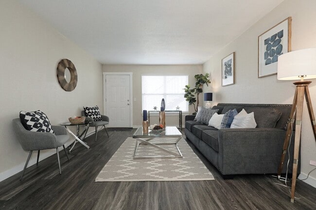 Living room with dark hardwood floors, gray sofa and accent chair, glass coffee table, patterned rug, and framed artwork