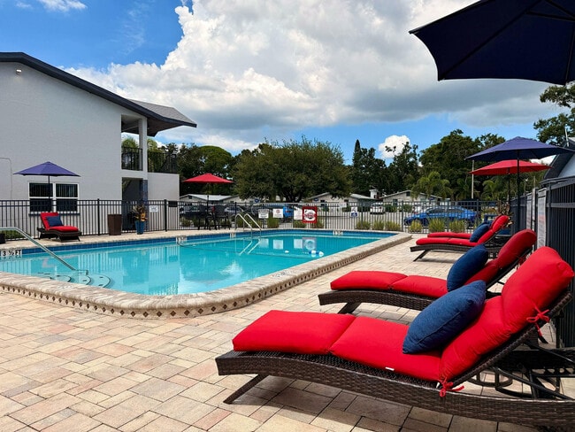 Inviting apartment complex pool area with bright red loungers and umbrellas under a clear blue sky.