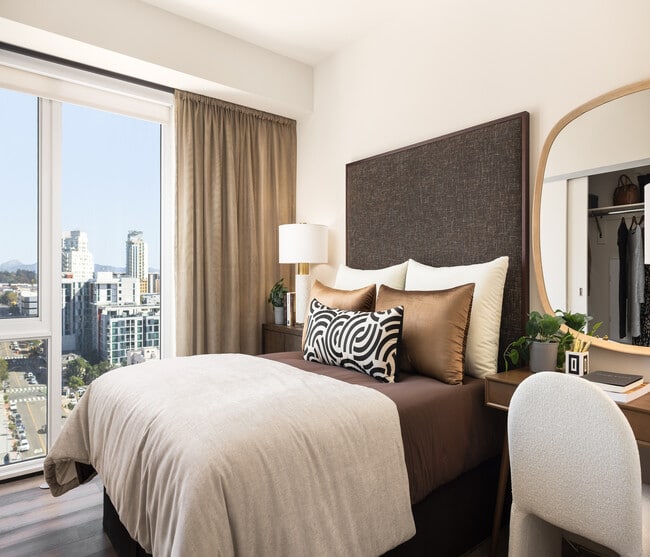 Chic and cozy bedroom featuring a plush bed with decorative pillows, elegant desk with mirror, and a stunning city view through large windows.