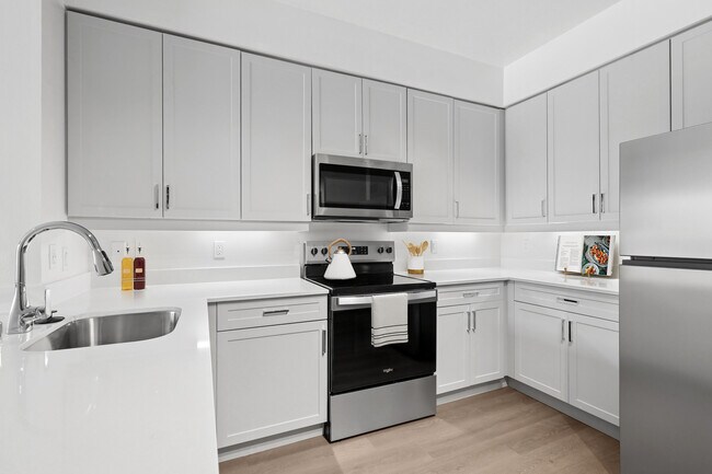Renovated Package II kitchen with stainless steel appliances, grey cabinetry, white quartz countertops, and hard surface flooring - Avalon at Cahill Park