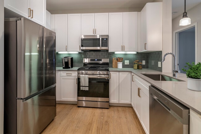 Renovated Package II kitchen with stainless steel appliances, grey quartz countertops, white cabinetry, grey tile backsplash, and hard surface flooring - Avalon at Assembly Row Apartments