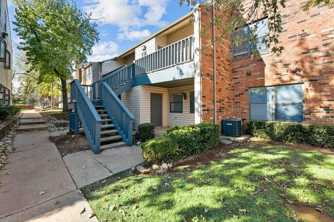 Two-story brick and siding apartment building with blue staircase, upper balcony, green lawn, and mature shade trees
