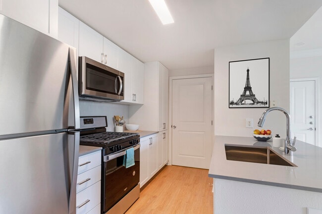 Finish Package II kitchen with white cabinetry, grey quartz countertops, extended hard surface flooring in select units, stainless steel appliances, and tile backsplash
