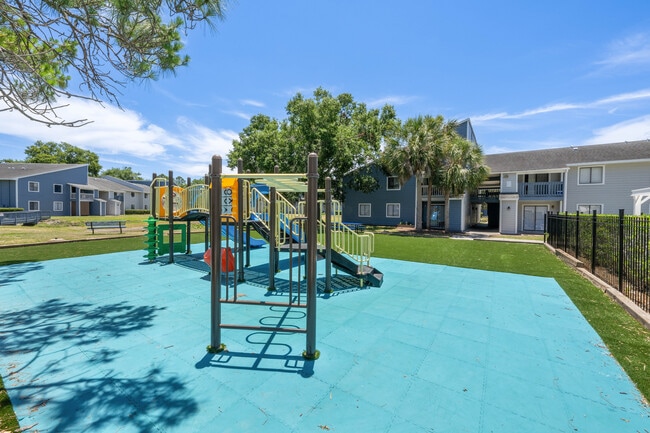 Un encantador parque infantil comunitario con juegos de colores vivos, rodeado de exuberante vegetación y modernos edificios de apartamentos bajo un cielo azul despejado. - Vista North
