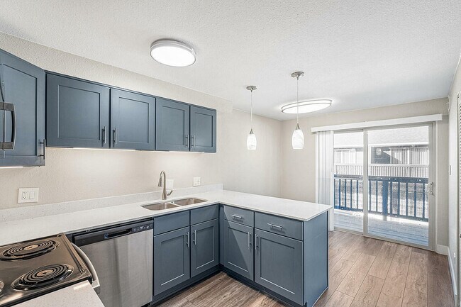 Modern kitchen with sleek blue cabinetry, stainless steel appliances, and a stylish pendant light over the countertop, adjacent to a bright living area with glass sliding doors to a balcony.