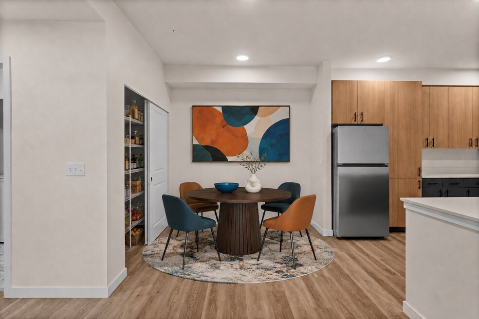 Staged dining room with a round table, four chairs, an area rug, abstract artwork, and a stocked open pantry.