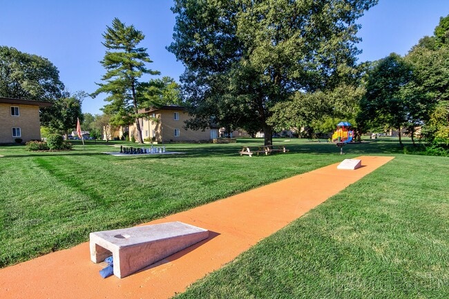 Community courtyard with two cornhole boards surrounded by green grass, picnic tables, and a playground.