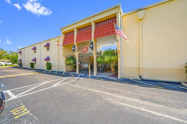Charming apartment complex with striking red tile roof, welcoming entrance, and lush palm landscaping under a bright blue sky.
