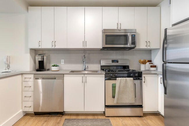Renovated II North/South kitchen with stainless steel appliances, grey quartz countertops, white lacquer cabinetry, white tile backsplash, and hard surface flooring