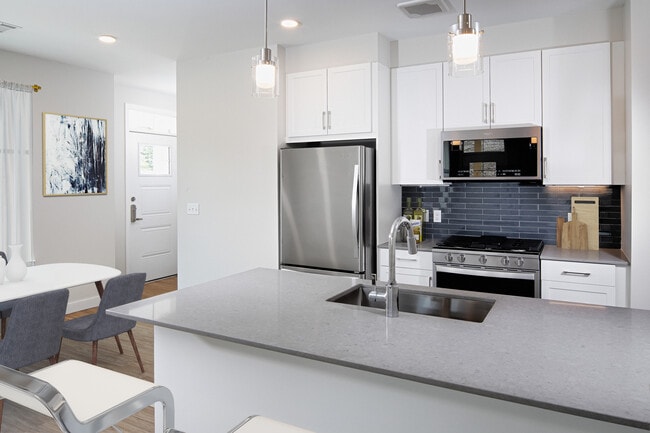 Phase II Renovated Package I apartment kitchen with stainless steel appliances, grey quartz countertops, white cabinetry, dark grey tile backsplash, and hard surface flooring - Avalon Acton