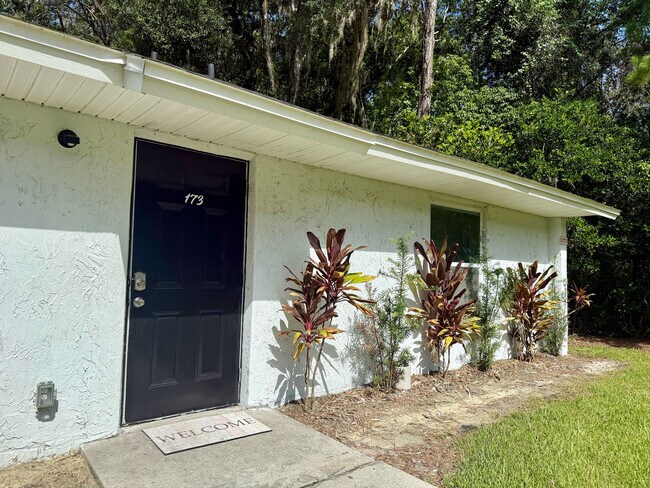 Charming home entrance featuring a dark door framed by lush greenery and vibrant plants, inviting atmosphere with a welcoming doormat. - Sunny Creek Apartments