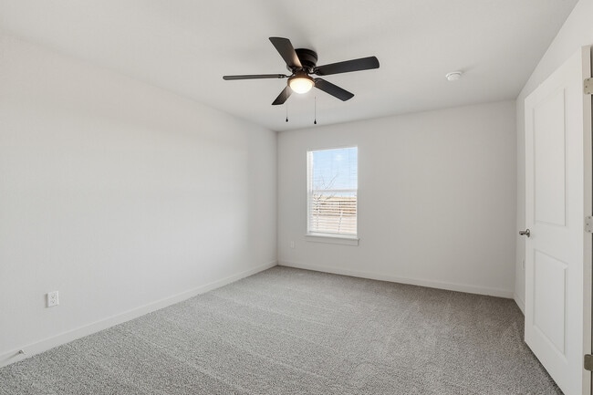 Dormitorio amplio y luminoso con lujosa alfombra gris, ventilador de techo moderno y ventana grande que brinda luz natural. - Oaks at Mustang