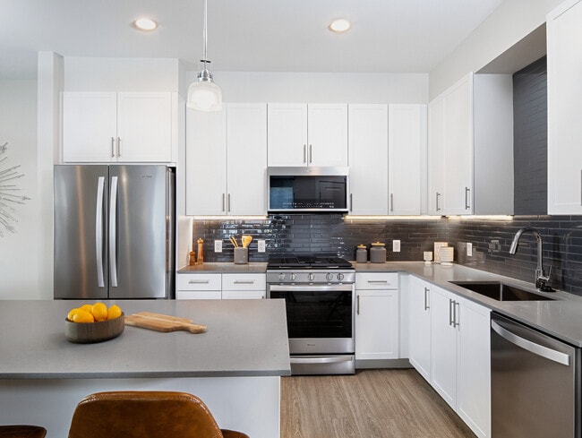 Renovated Package II kitchen with white cabinetry, grey quartz countertops, grey tile backsplash, stainless steel appliances, and hard surface plank flooring