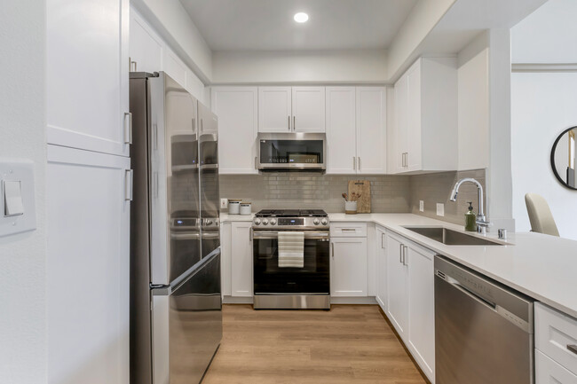 Renovated Package I kitchen with stainless steel appliances, white speckled quartz countertop, white cabinetry, grey tile backsplash, and hard surface flooring - Avalon Thousand Oaks Plaza Apartments