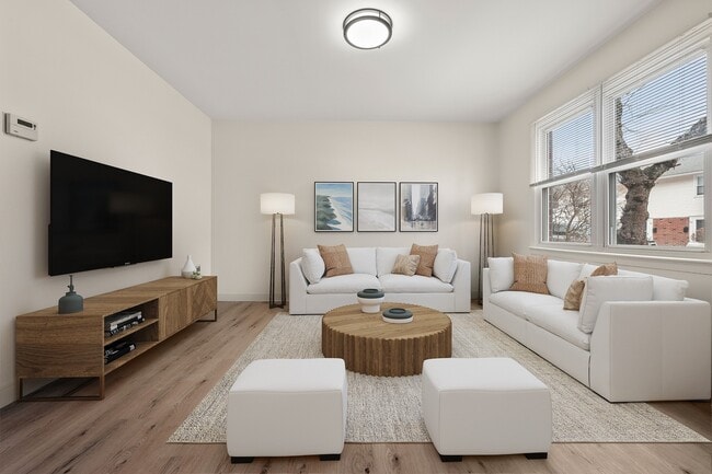 Bright and inviting apartment living room with modern decor, featuring large windows, cozy white sofas, and a stylish wooden coffee table, perfect for relaxation and entertainment. - Stony Brook Commons