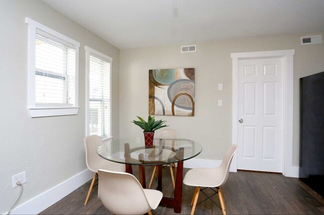 Bright dining area with round glass table, white modern chairs, hardwood floors, and two windows with blinds