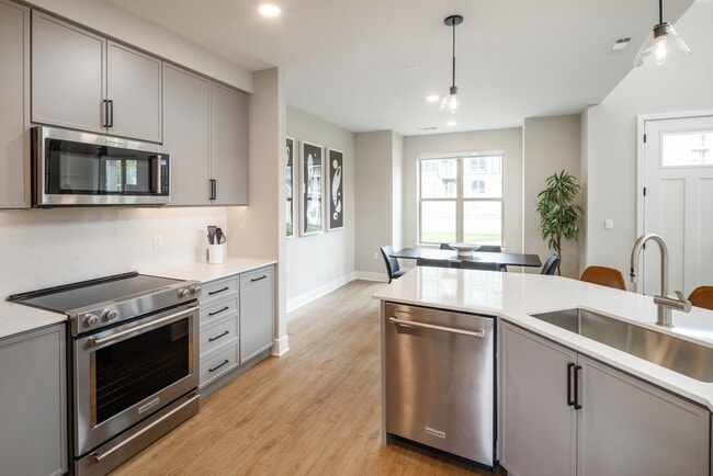 Signature Collection Townhome kitchen with upgraded stainless steel appliances, white quartz countertops and backsplash, grey cabinetry, hard surface flooring, and pendant lighting. - Avalon Wayne
